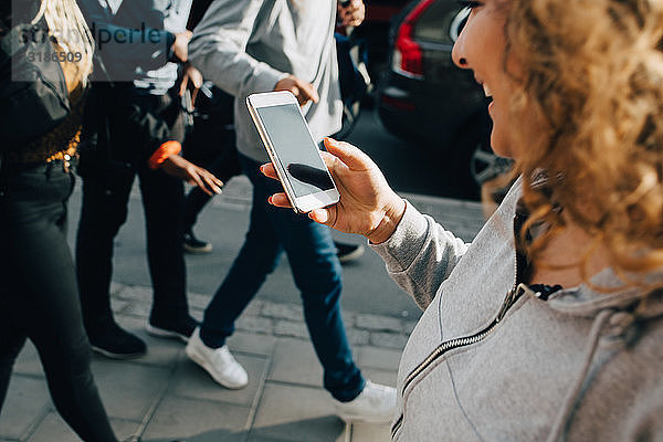 Junge Frau benutzt Mobiltelefon beim Gehen mit Freunden auf dem Bürgersteig