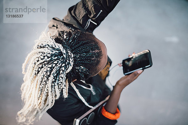 Schrägaufnahme eines Teenager-Mädchens mit Dreadlocks  das tanzt  während es sein Smartphone im Skateboard-Park in der Hand hält