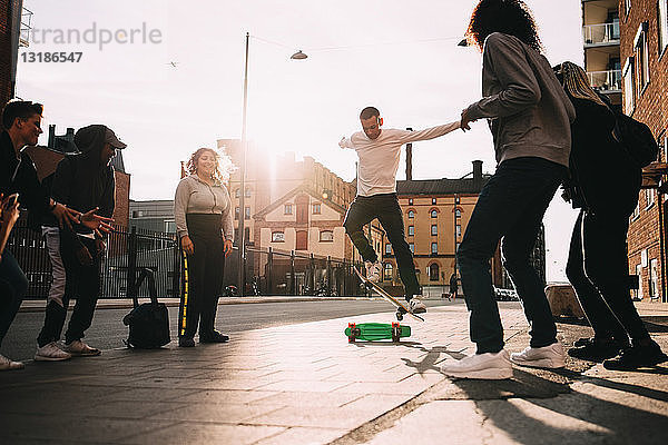 Freundinnen und Freunde schauen einem jungen Mann zu  der in der Stadt einen Stunt auf dem Skateboard macht