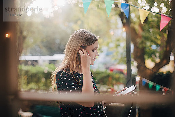 Junge Frau beim Anpassen von Kopfhörern während der Benutzung eines Mobiltelefons auf dem Balkon während einer Party
