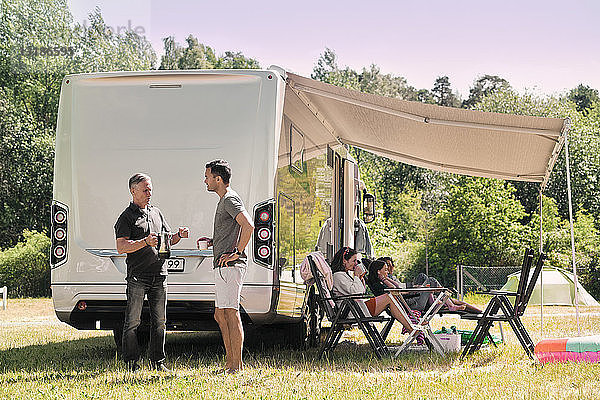 Älterer Mann unterhält sich mit seinem Sohn  während sich die Familie vor dem Lieferwagen auf dem Campingplatz entspannt