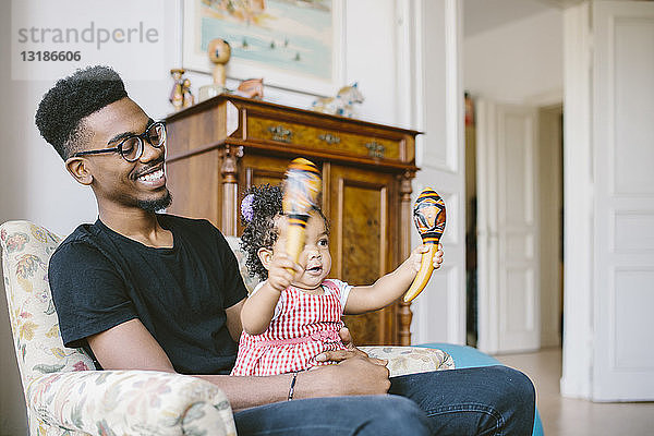 Kleines Mädchen hält Maracas  während es zu Hause mit dem glücklichen Vater auf einem Sessel sitzt