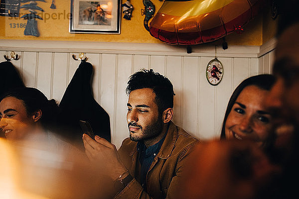 Junger Mann benutzt Mobiltelefon  während er inmitten von Freunden im Restaurant sitzt