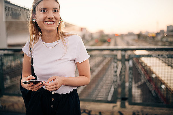 Porträt einer lächelnden jungen Frau  die ein Smartphone in der Hand hält  während sie auf einer Brücke in der Stadt steht