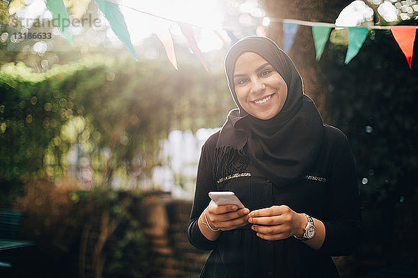 Porträt einer lächelnden Frau im Hijab mit einem Smartphone in der Hand im Hinterhof