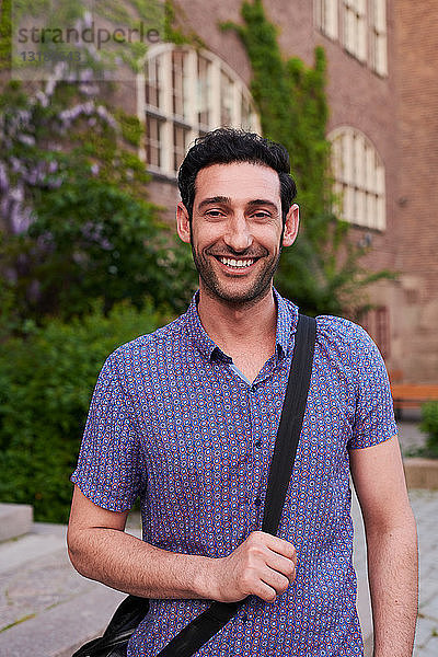 Porträt eines lächelnden jungen Mannes mit Umhängetasche vor der Sprachschule stehend