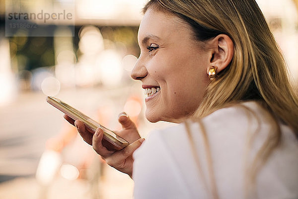Lächelnde junge Frau hält Handy in der Hand  während sie draußen in der Stadt sitzt