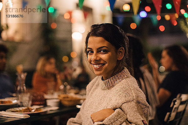 Porträt einer lächelnden jungen Frau  die während einer Dinnerparty im Hinterhof am Tisch sitzt
