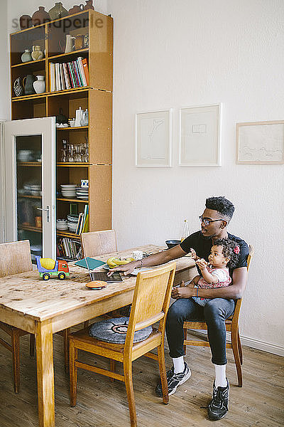 Junger Mann benutzt Laptop  während er mit seiner Tochter auf einem Stuhl am Esstisch im Haus sitzt