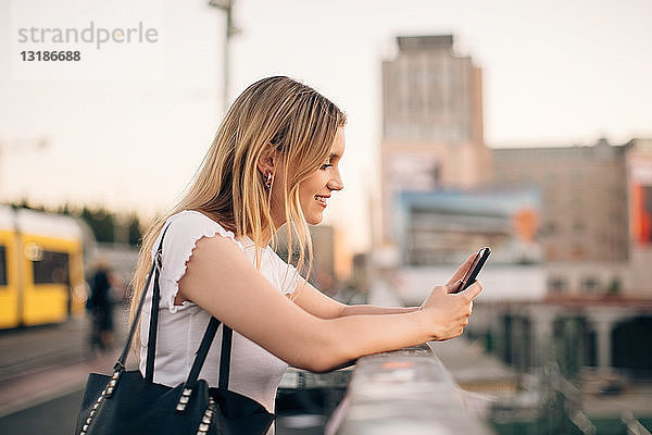 Seitenansicht einer jungen Frau  die ein Mobiltelefon benutzt  während sie sich an das Geländer einer Brücke in der Stadt lehnt