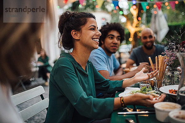 Lächelnde junge Frauen gestikulieren  während sie während der Dinnerparty mit Freunden am Tisch sitzen