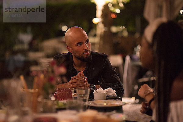 Junger Mann mit kahlrasiertem Kopf schaut eine Freundin an  während er während der Dinnerparty am Tisch sitzt