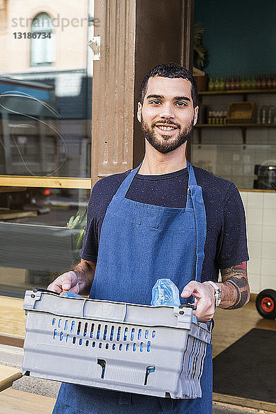 Porträt eines lächelnden jungen männlichen Besitzers  der eine Kiste gegen ein Restaurant trägt