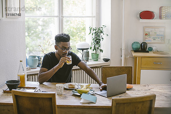 Junger Mann trinkt Saft  während er am Tisch im Haus auf seinen Laptop schaut