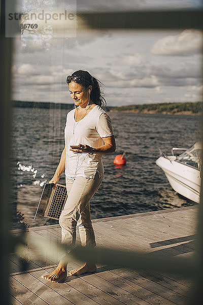 Frau in voller Länge mit Mobiltelefon und Laptop auf der Terrasse am See