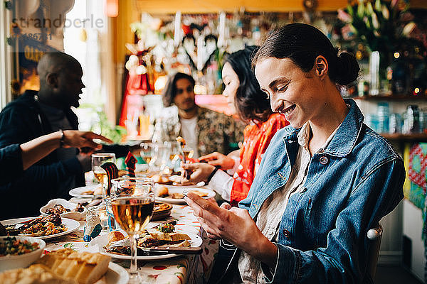 Lächelnde junge Frau  die ein Smartphone benutzt  während sie während einer Dinnerparty mit Freunden im Restaurant am Tisch sitzt