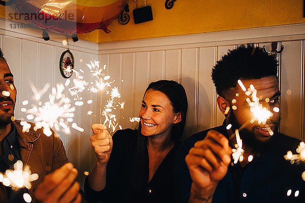 Lächelnde junge Frau mit multiethnischen männlichen Freunden  die während der Dinnerparty im Restaurant Wunderkerzen halten