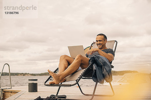 Lächelnder reifer Mann mit Laptop  während er auf einem Liegestuhl am Steg vor bewölktem Himmel sitzt