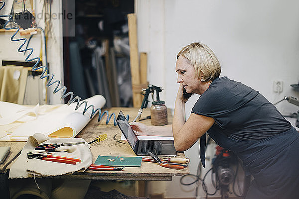 Seitenansicht einer reifen Polsterin  die während eines Telefongesprächs in der Werkstatt auf den Laptop schaut