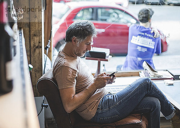 Erwachsener Besitzer benutzt Mobiltelefon  während er im Feinkostladen auf einem Stuhl sitzt