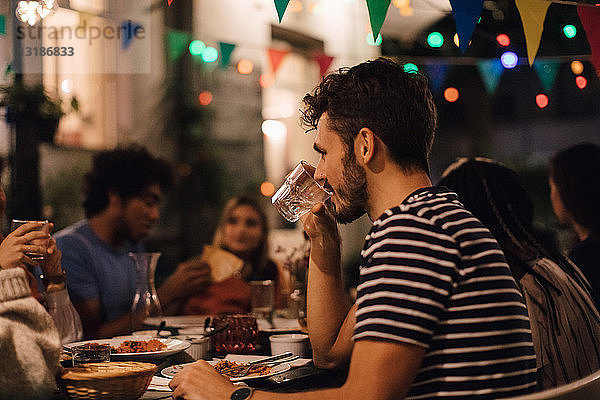 Junger Mann trinkt Wasser  während er während einer Gartenparty mit Freunden zu Abend isst