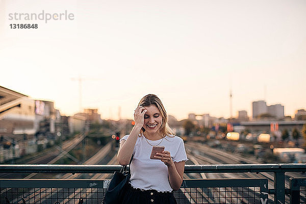 Lächelnde junge Frau benutzt Smartphone auf der Brücke gegen den klaren Himmel während des Sonnenuntergangs in der Stadt