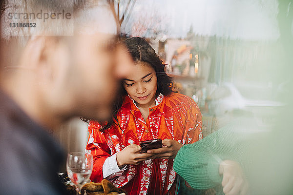 Junge Frau benutzt ein Smartphone  während sie mit Freunden im Restaurant sitzt und durch ein Glasfenster gesehen wird