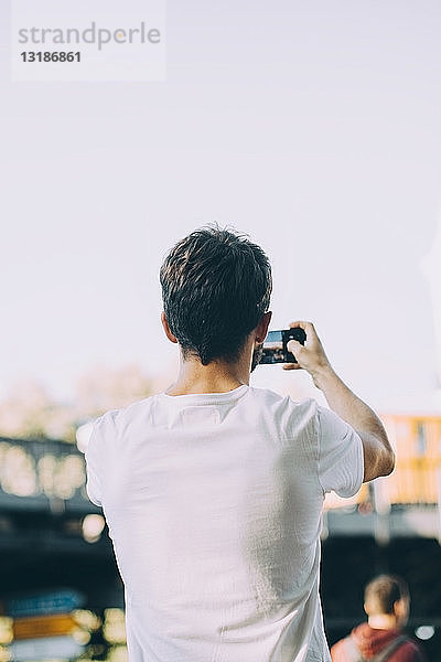 Rückansicht eines jungen Mannes  der auf der Straße stehend per Handy fotografiert