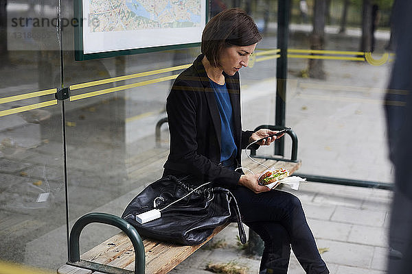 Mittlere erwachsene Geschäftsfrau benutzt ein Smartphone  während sie an einer Bushaltestelle in der Stadt ein Sandwich isst