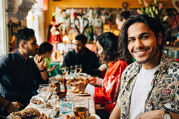 Porträt eines lächelnden jungen Mannes  der mit multi-ethnischen Freunden zusammensitzt und eine Dinnerparty im Restaurant genießt