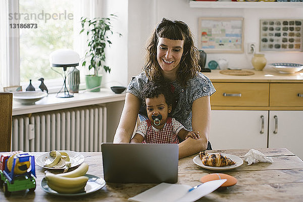 Lächelnde Frau mit Laptop  während sie mit ihrer Tochter am Esstisch im Haus sitzt