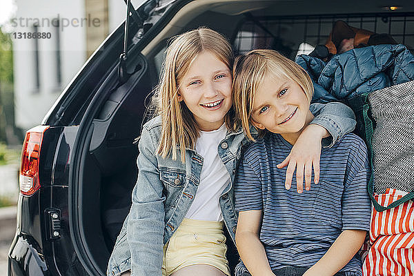 Porträt eines lächelnden blonden Mädchens  das mit dem Arm um die Schwester im Kofferraum eines Autos vor dem Haus sitzt