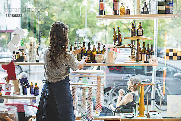 Rückansicht einer weiblichen Mitarbeiterin  die im Feinkostladen Flaschen im Regal arrangiert