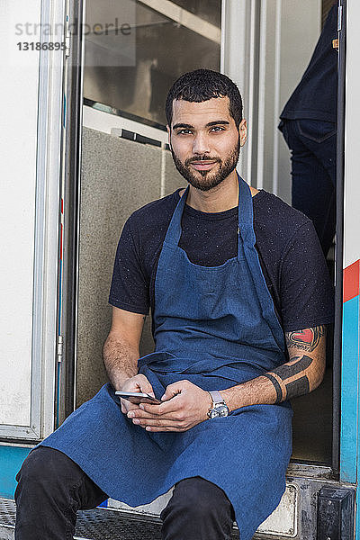 Porträt eines selbstbewussten jungen Unternehmers  der mit einem Smartphone am Eingang eines Speisewagens sitzt