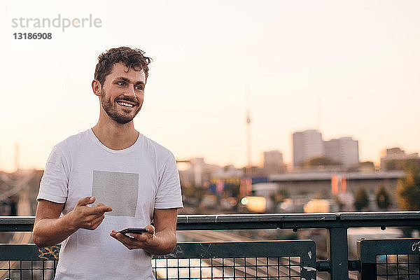 Lächelnder junger Mann schaut weg  während er bei Sonnenuntergang sein Handy auf der Brücke gegen den klaren Himmel hält