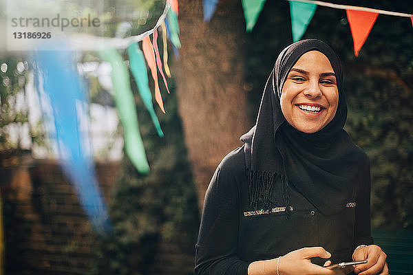 Porträt einer lächelnden jungen Frau im Hijab  die im Hinterhof ein Mobiltelefon in der Hand hält