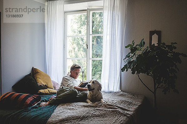 Ein Mann im Ruhestand liest in voller Länge ein Buch  während er sich mit seinem Hund zu Hause im Bett ausruht