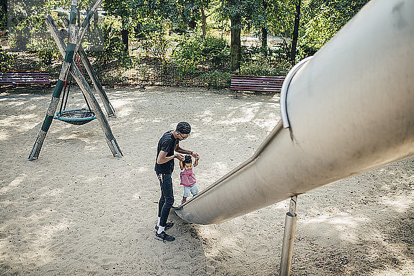 Vater hält Hände einer Tochter  die auf einer Rutsche auf einem Spielplatz läuft