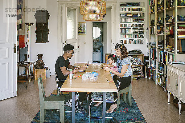 Junger Mann benutzt Laptop  während Frau mit Tochter am Esstisch im Haus spielt