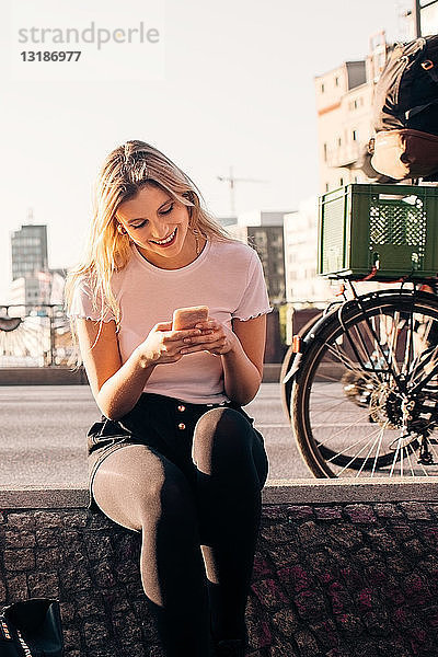 Lächelnde junge Frau benutzt Mobiltelefon  während sie auf einer Stützmauer an einer Straße in der Stadt sitzt