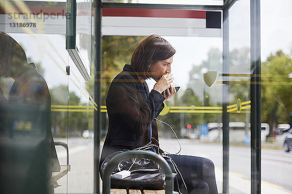 Seitenansicht einer Geschäftsfrau  die ein Sandwich isst  während sie an einer Bushaltestelle in der Stadt wartet