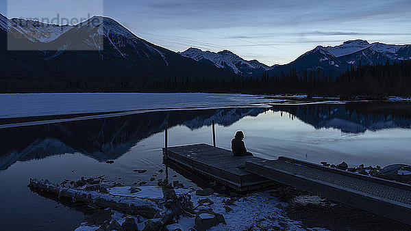 Frau sitzt an einem ruhigen Wintersee mit Blick auf die Berge bei Nacht  Banff  Alberta  Kanada
