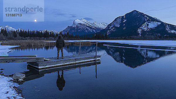 Frau an einem ruhigen Winterseesteg mit Blick auf den Vollmond über den Bergen  Banff  Alberta  Kanada