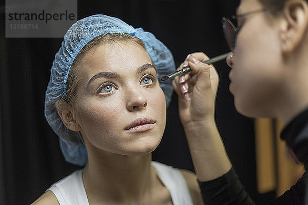 Visagistin trägt Make-up auf die Augen einer jungen Frau mit Duschhaube auf