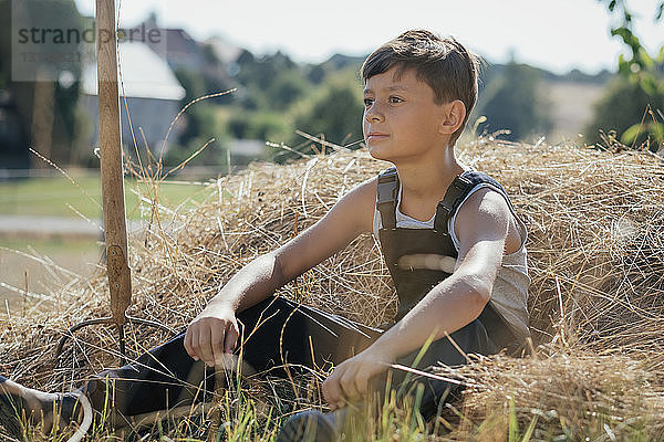 Junge in Latzhose sitzt im Heu auf einem sonnigen Bauernhof