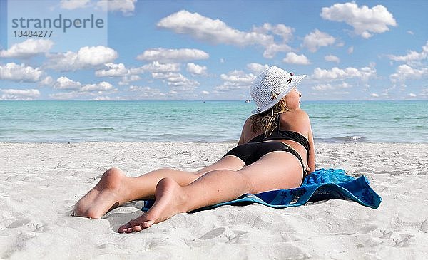 Mädchen mit Hut an einem weißen Sandstrand mit türkisfarbenem Meer auf einem blauen Handtuch liegend