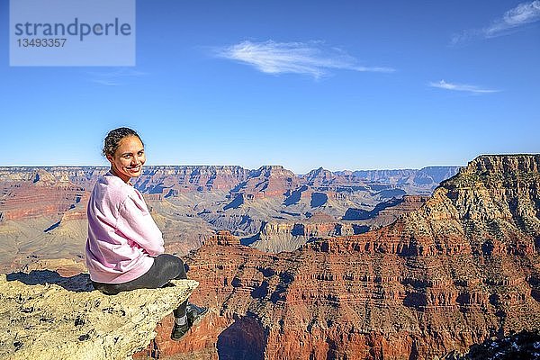 Junge lächelnde Frau  sitzend am Abgrund der gigantischen Schlucht des Grand Canyon  Blick vom Rim Trail  zwischen Mather Point und Yavapai Point  erodierte Felslandschaft  South Rim  Grand Canyon National Park  nahe Tusayan  Arizona  USA  Nordamerika