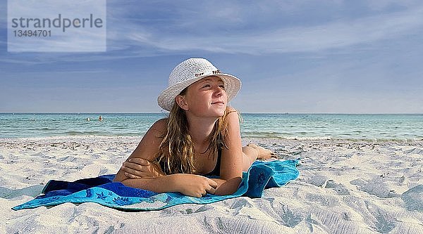 Mädchen mit Sonnenhut auf einem blauen Strandtuch am Meer an einem weißen Sandstrand liegend
