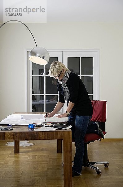 Eine Architektin kniet auf einem Stuhl am Schreibtisch in ihrem Büro und skizziert einen Plan