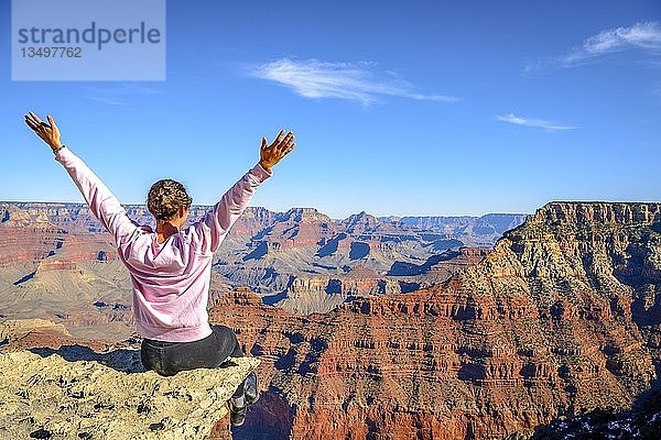 Junge Frau  die mit ausgestreckten Armen glücklich in die Ferne schaut  sitzend am Abgrund der gigantischen Schlucht des Grand Canyon  Blick vom Rim Trail  zwischen Mather Point und Yavapai Point  erodierte Felslandschaft  South Rim  bei Tusayan  Arizona  USA  Nordamerika
